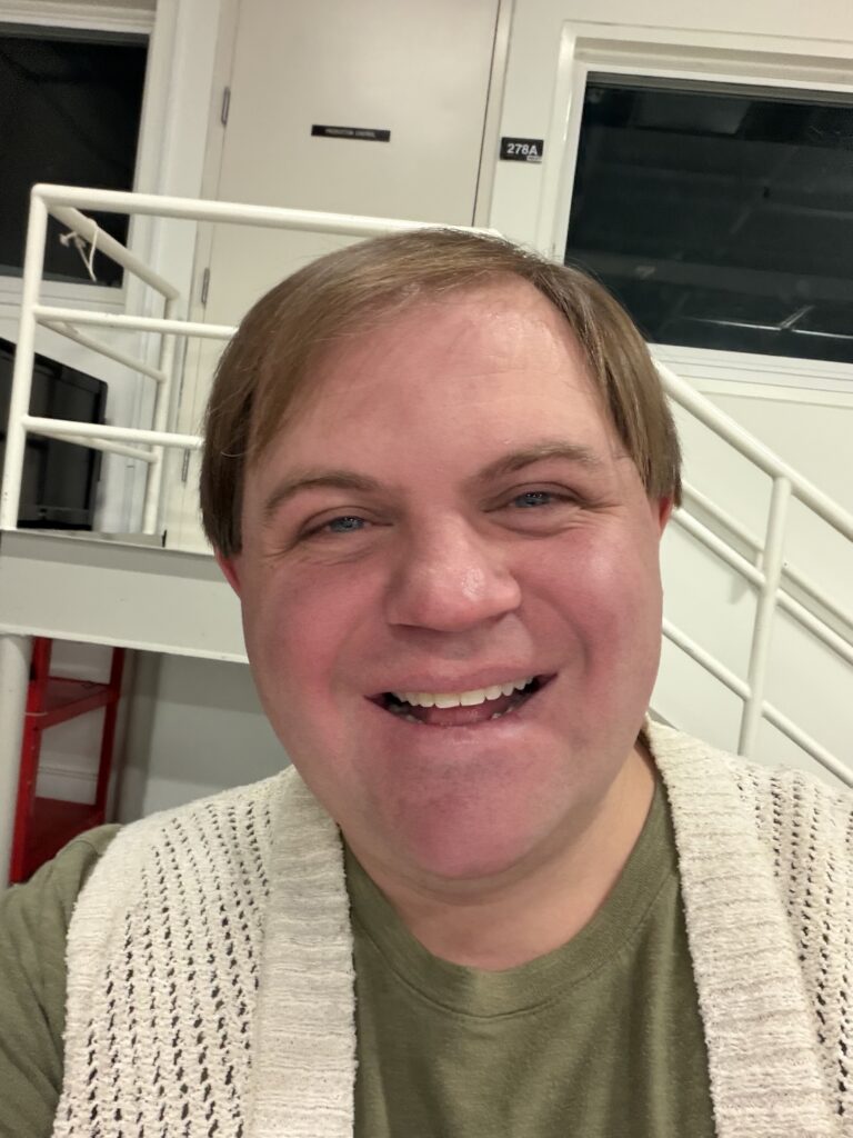 An image of Cody Mejeur wearing a green shit and white knitted vest. Taken at Global Game Jam 2026.